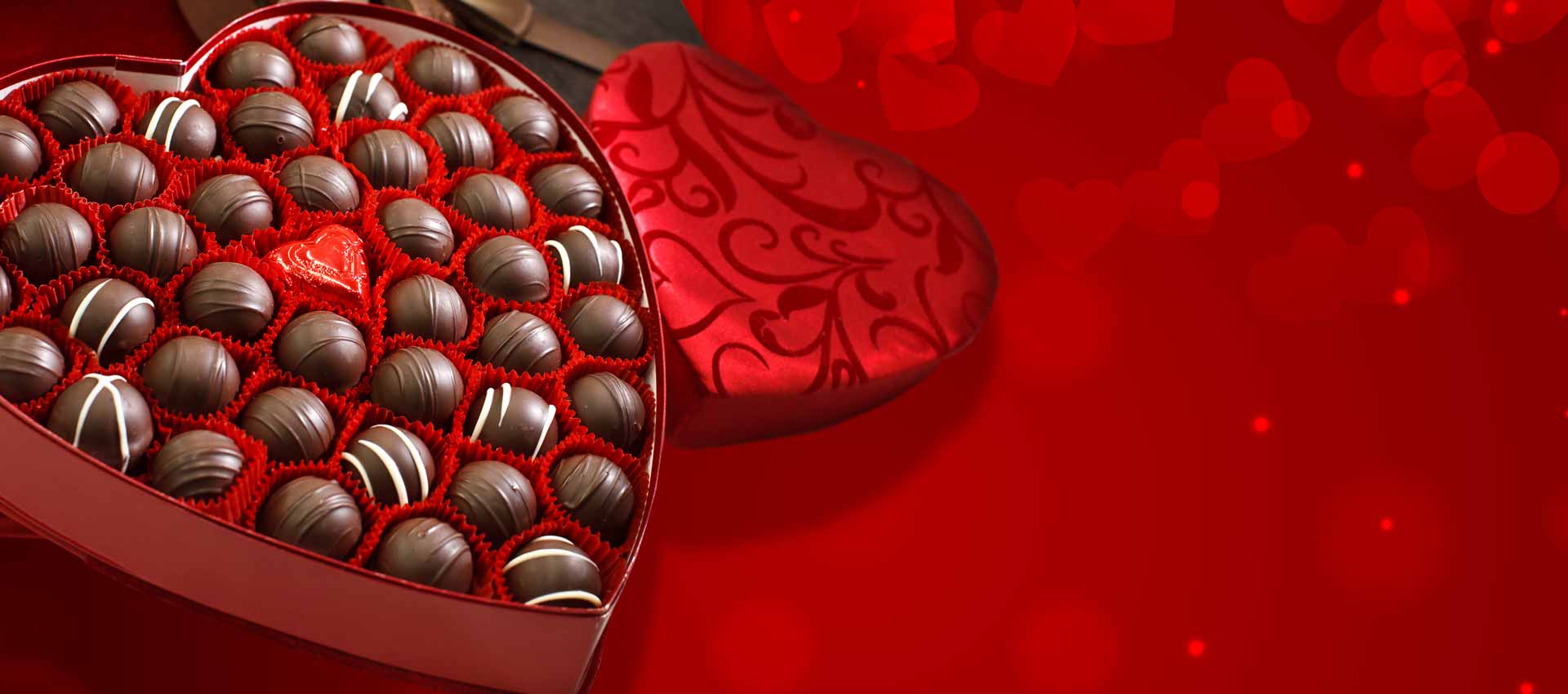 An open heart box of chocolate truffles with a red background of hearts and heart boxes