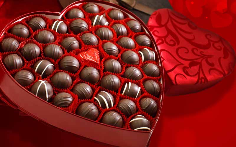 An open heart box of chocolate truffles with a red background of hearts and heart boxes