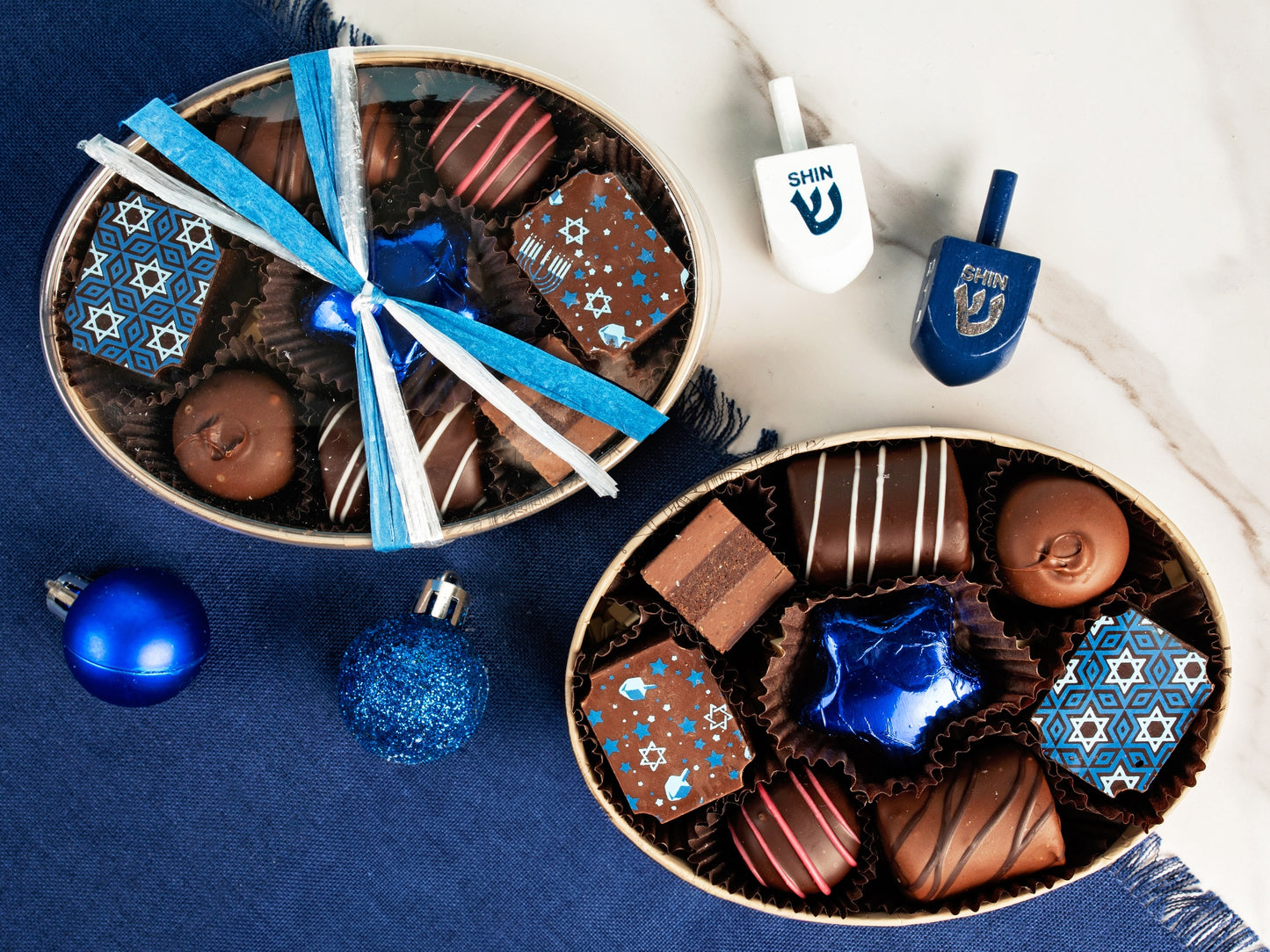 An oval box filled with Chanukah chocolates, next to the same box with a clear lid and blue and white ribbon.