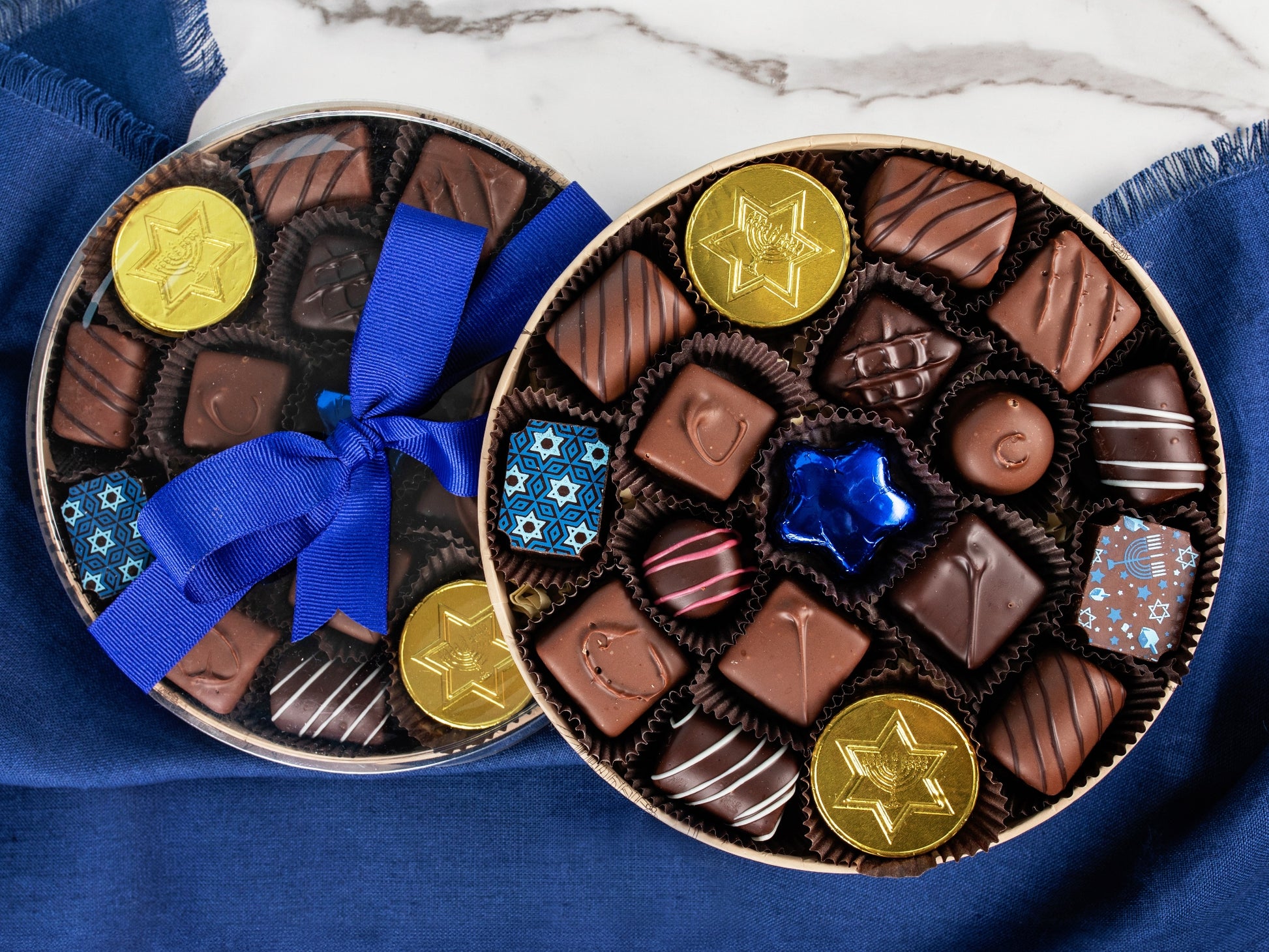 An open round box filled with chocolates next to a closed box with a clear lid and blue ribbon.