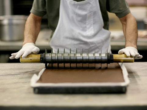 a man wearing white gloves rolling a cutting tool across a tray of fudge.