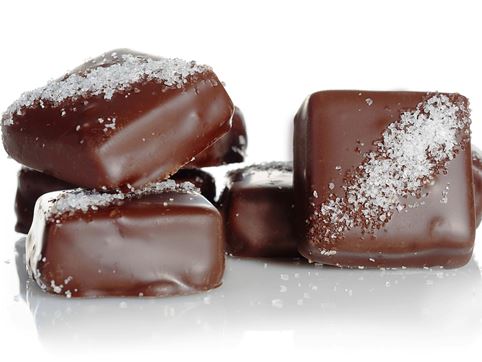 Caramel squares enrobed in milk and dark chocolate are stacked up together. The squares have a diagonal line of sea salt across the top.  